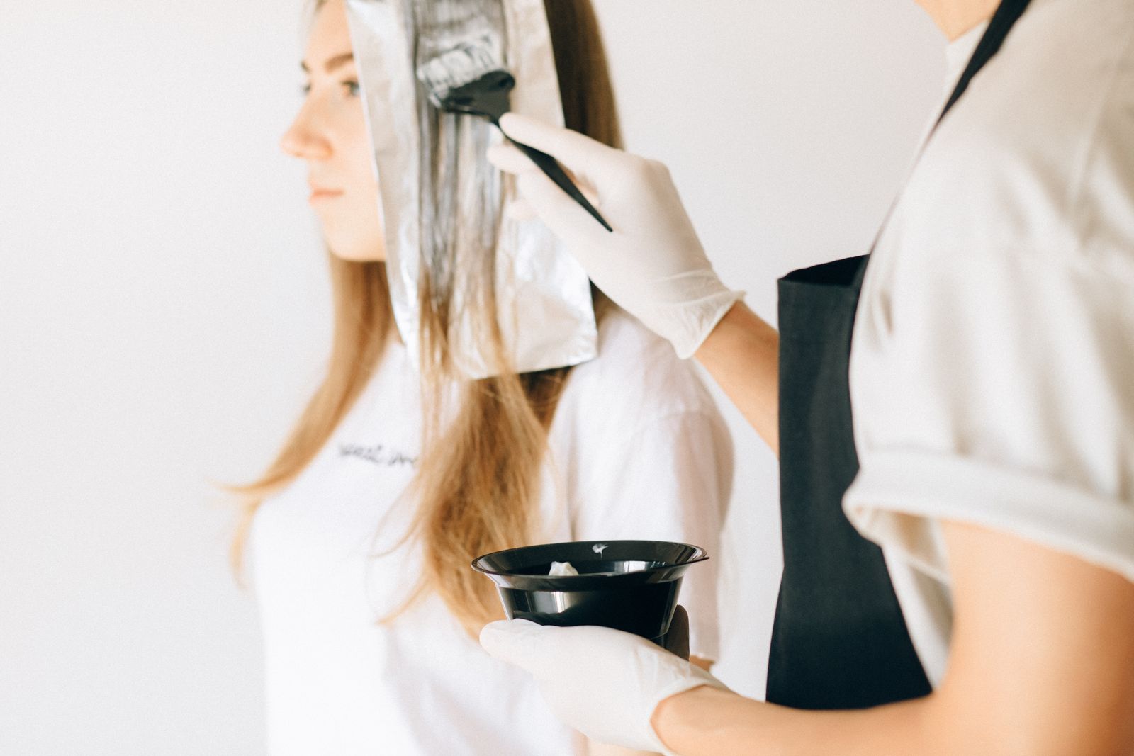 A stylist in gloves painting color onto a client’s hair from a black bowl