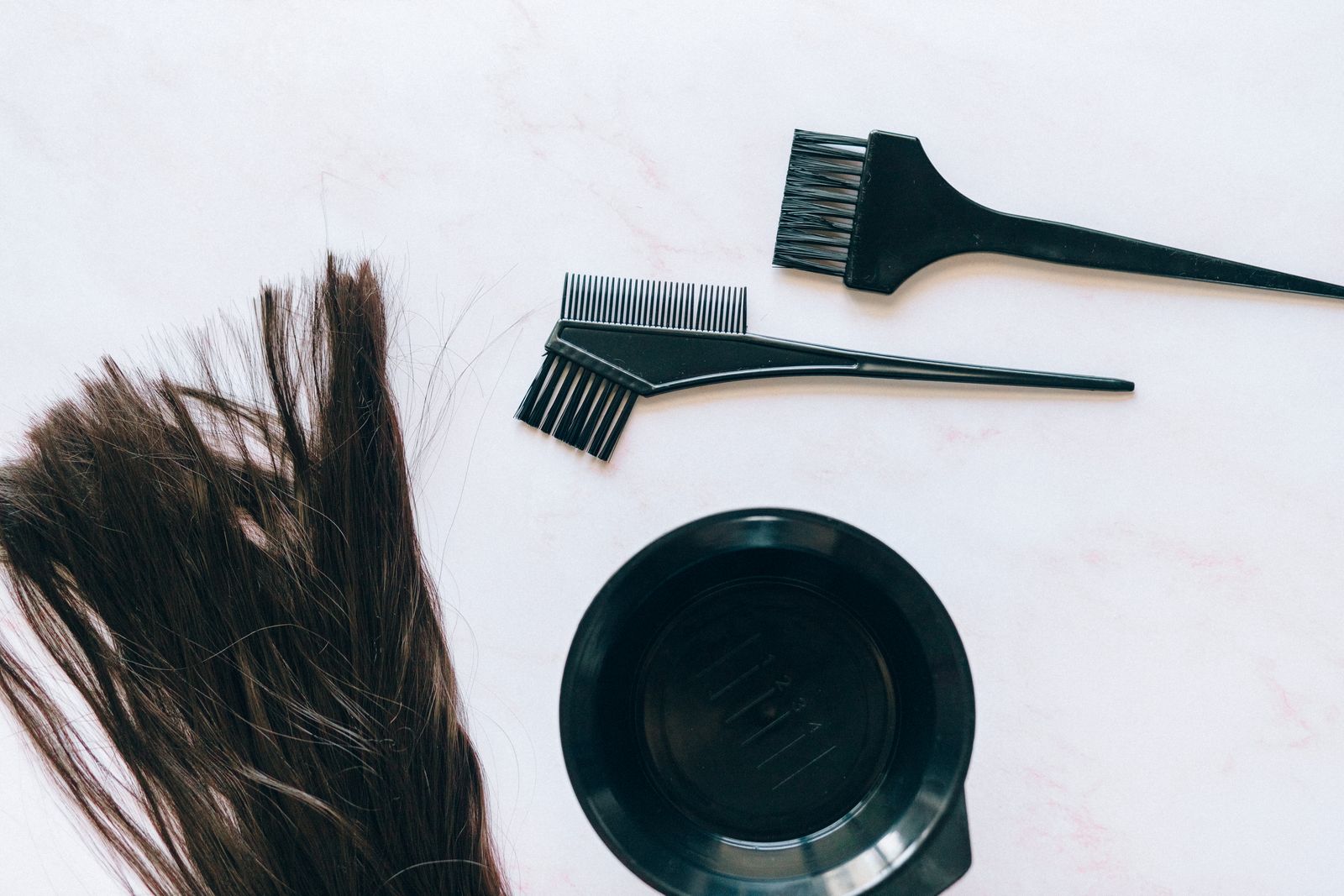 Hair color brushes, a mixing bowl and a lock of brown hair on a white surface