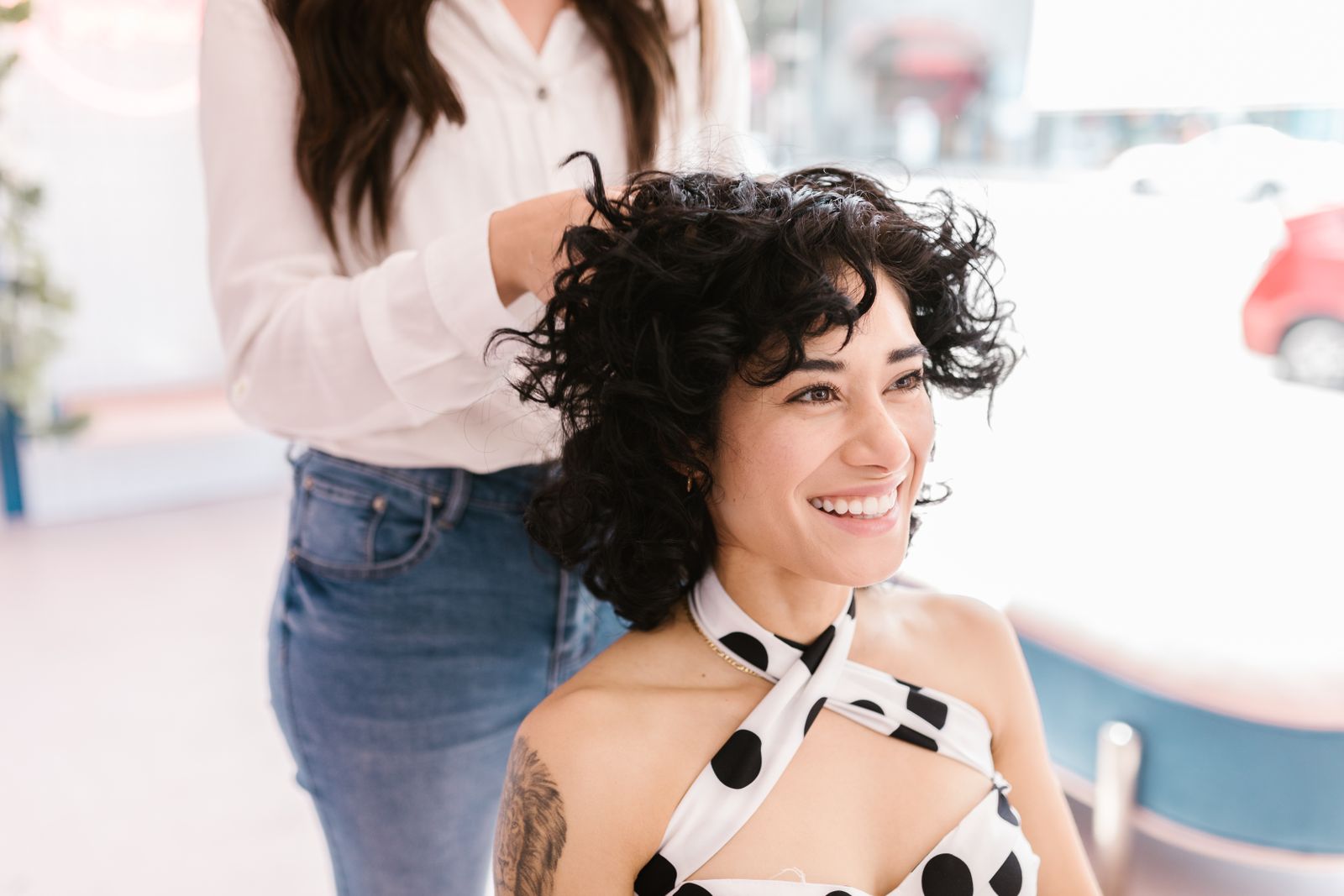 A smiling client with dark curls being styled by a stylist in a bright salon