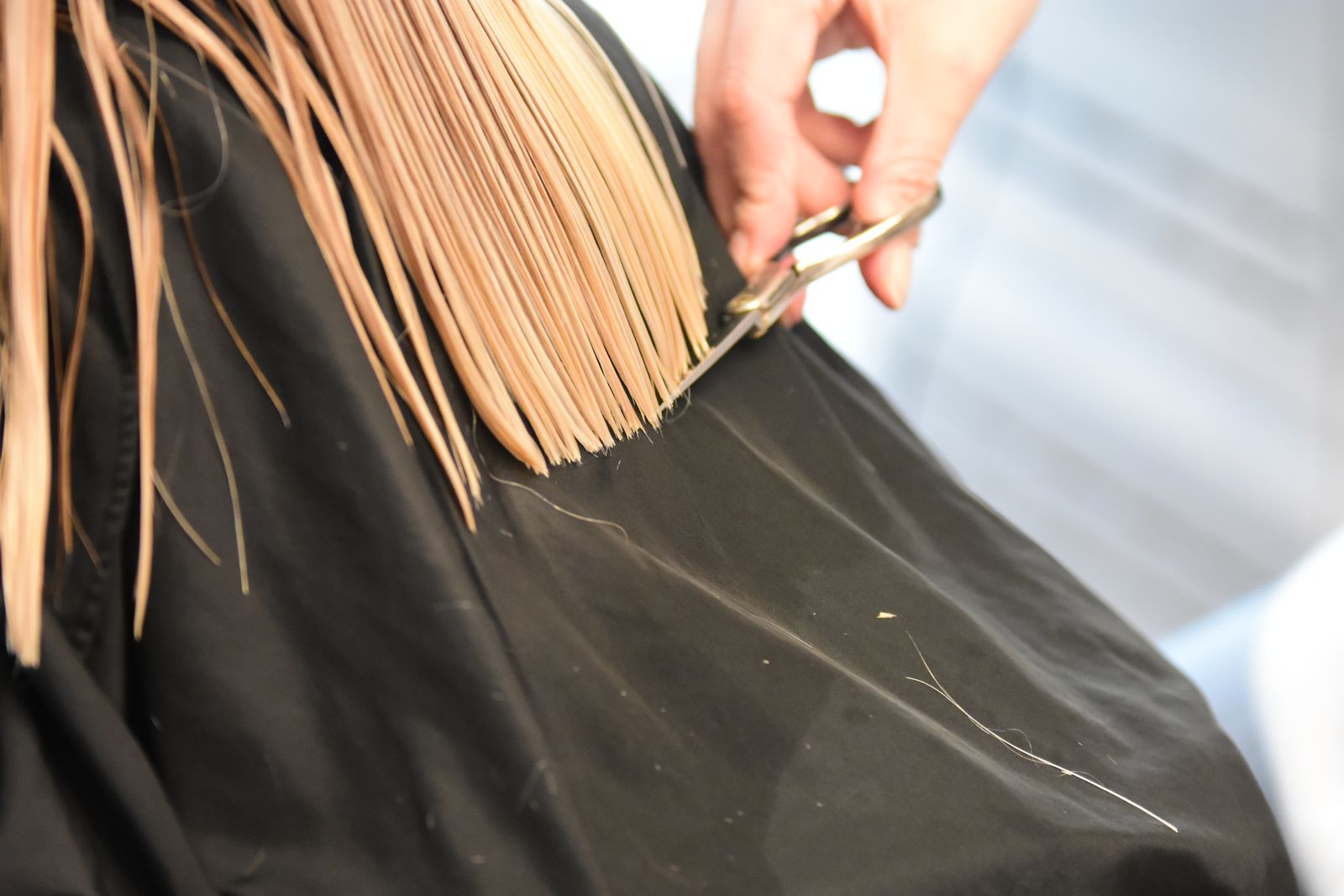 A stylist trimming long blonde hair with shears over a black cape