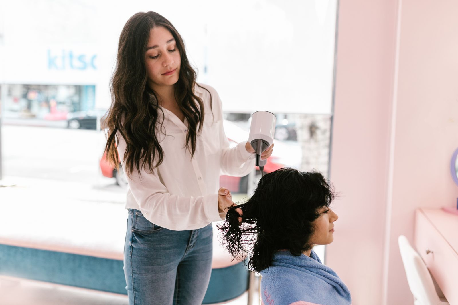 A stylist in a white blouse drying a client’s dark wavy hair